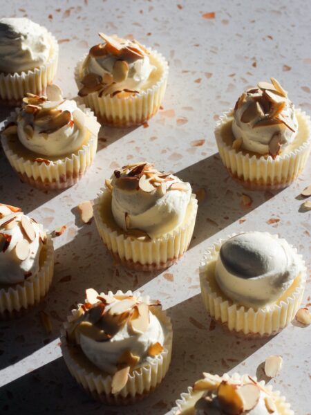 Mini almond cheesecakes topped with whipped cream and toasted sliced almonds, photographed in warm natural sunlight.