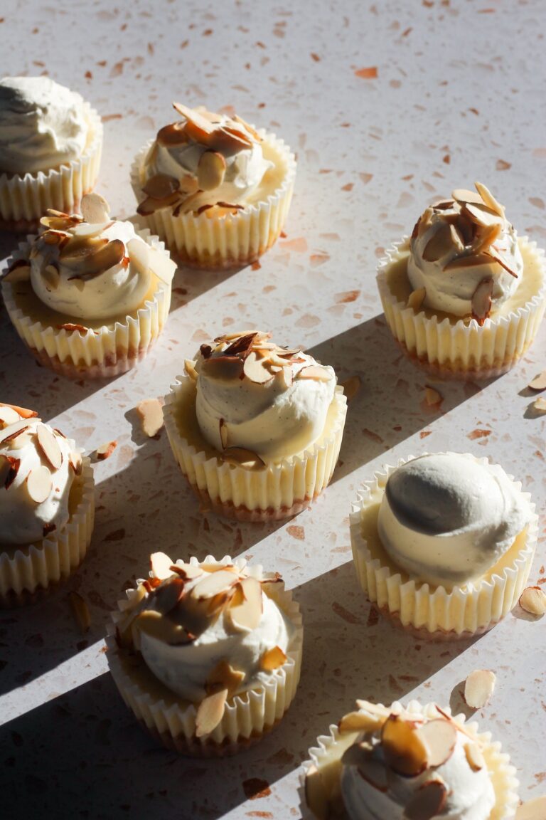 Mini almond cheesecakes topped with whipped cream and toasted sliced almonds, photographed in warm natural sunlight.